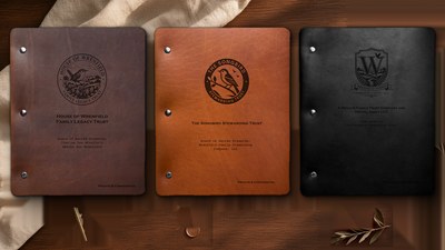 Three House of Wrenfield folios in a row — deep chocolate brown, cognac brown and matte black leather, each embossed with a small circular seal, staged on a dark walnut table in warm golden light